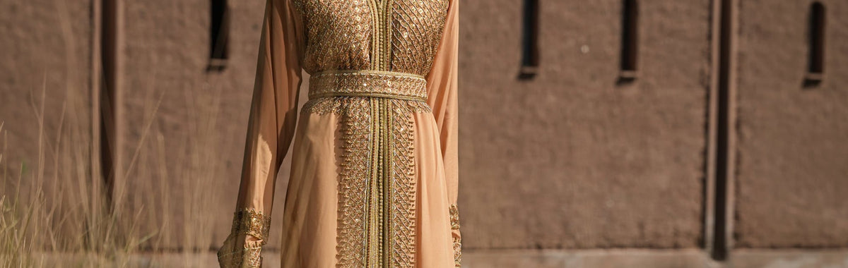 Apricot crepe Moroccan kaftan with a gold sequin plastron and embroidered belt, against a Marrakech adobe wall